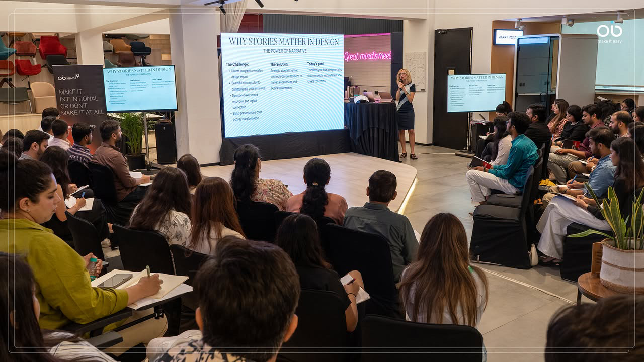 A diverse audience of approximately 30 attendees seated on the floor and cushions, facing a female presenter standing at a podium in front of projection screens displaying 'Why Stories Matter in Design' presentation slides in a modern interior design studio space.